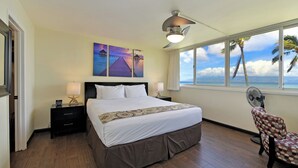 Ein Hotelzimmer mit einem Bett, zwei Nachttischen mit Lampen, einem Deckenventilator, einem Fenster mit Blick auf Palmen und das Meer sowie einem Stuhl mit Punkten.