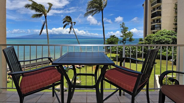 Ein Balkon mit Tisch und Stühlen, der Blick auf ein klares, blaues Meer und Palmen bietet, mit einem Gebäude im Hintergrund.