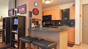 A kitchen with wooden cabinets, a black microwave, and a small island with stools. There are clocks on the wall and a guitar in the corner. - Ocean View Condo in Kahana - Valley Isle 302 (Lahaina)