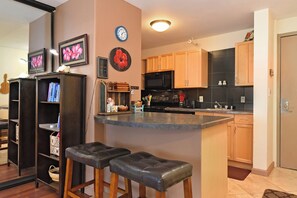 A kitchen with wooden cabinets, a black microwave, and a small island with stools. There are clocks on the wall and a guitar in the corner. - Ocean View Condo in Kahana - Valley Isle 302 (Lahaina)