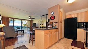 A modern kitchen with wooden cabinets, a black refrigerator, and a dining area with a view of the ocean. - Ocean View Condo in Kahana - Valley Isle 302 (Lahaina)