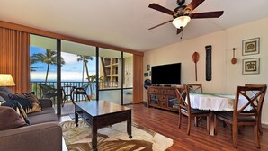 A living room with a large window, a ceiling fan, a flat-screen TV, and a dining area with a table and chairs. - Ocean View Condo in Kahana - Valley Isle 302 (Lahaina)