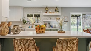 Une cuisine moderne avec des meubles blancs, un îlot central doté de chaises en osier et une vue sur l’extérieur.