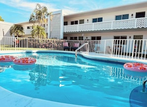 A swimming pool with a white fence, palm trees, and a building with balconies. - Fernando Flats: Boutique Studio Beach Flat 11 (Palm Beach Shores)