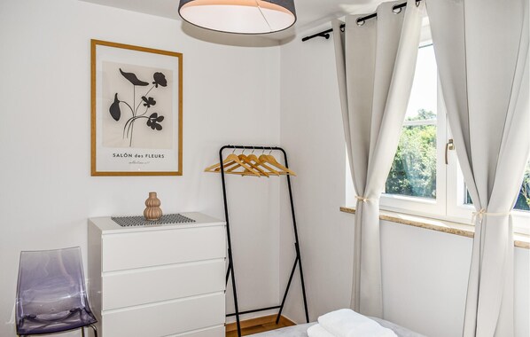 Une chambre à coucher comprenant un lit, une commode, une chaise, un tableau encadré, un étendoir et une fenêtre avec des rideaux.