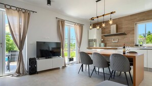 Une cuisine moderne dotée d’un grand îlot central, d’un coin repas avec quatre chaises, d’un téléviseur à écran plat et d’une vue sur la verdure environnante.