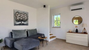 Un salon moderne avec un canapé gris, une petite table d’appoint avec une bouilloire, une commode blanche, un miroir rond et un tableau représentant un arbre.