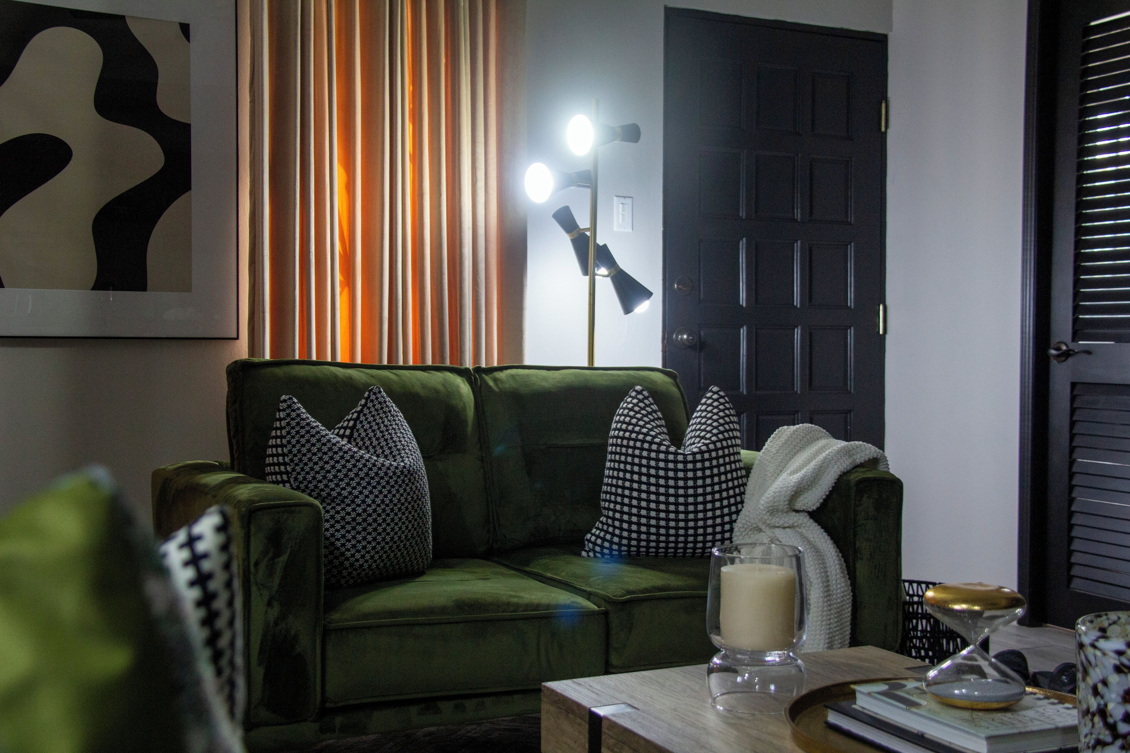 A green sofa with patterned pillows, a table with a candle and book, and a floor lamp.