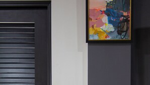 A door with horizontal slats, a framed abstract painting on the wall, and a potted plant with large green leaves. - Villa Torre II (Belize City)