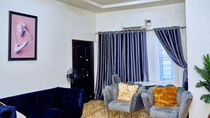Ein modernes Wohnzimmer mit einem dunkelblauen Sofa, einem grauen Sessel mit flauschigem Kissen, einem gerahmten Bild an der Wand und einem Deckenventilator.