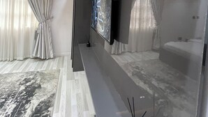 Ein modernes Wohnzimmer mit einem Flachbildfernseher an einer dunklen Wand, einer Zimmerpflanze auf einem Regal und einem großen, gemusterten Teppich auf dem Boden.