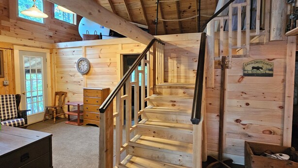 A wooden cabin interior with a staircase leading to a loft area, a dining table, and a clock on the wall. - Spacious cabin on beautiful Long Lake w/waterfront view, private dock, fire pit. (Princeton)