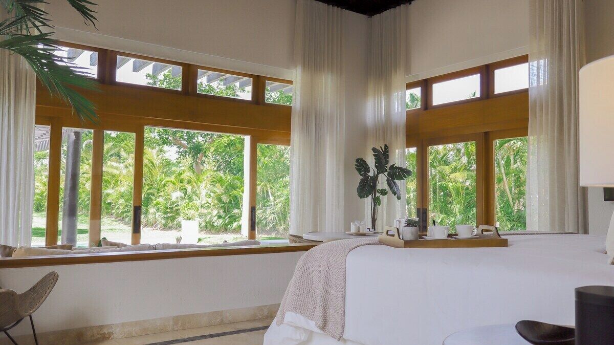 Un dormitorio amplio con ventanas grandes, una cama, una silla y una mesita de noche con una planta.