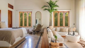 A spacious bedroom with a large bed, a sofa, a chair, and a coffee table. There is a large window with a view of greenery outside.