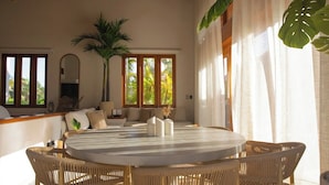 A spacious living area with a round table, wicker chairs, a sofa, and large windows with curtains.