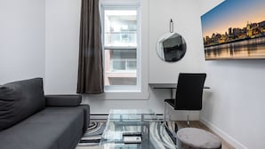 A modern living room with a grey sofa, a glass coffee table, a black chair, a round mirror, and a cityscape painting on the wall.
