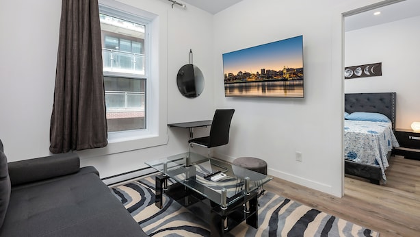 A modern living room with a glass coffee table, a sofa, a bed with a blue patterned bedspread, and a cityscape painting on the wall.