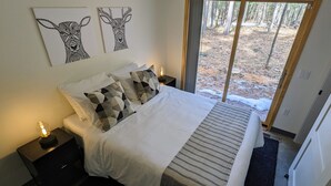 A bedroom with a large bed, two nightstands, a lamp, and two wall art pieces of deer. - Deer Cabin @ Orange Tractor Retreat: a new, 3BR modern cabin on 40 private acres (Wisconsin Dells)
