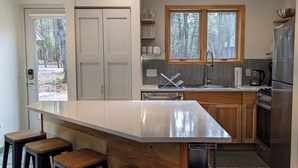A modern kitchen with a central island, wooden cabinets, a built-in oven, and a window overlooking a forest. - Deer Cabin @ Orange Tractor Retreat: a new, 3BR modern cabin on 40 private acres (Wisconsin Dells)