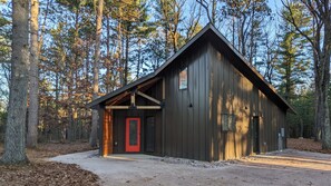 A cabin with a red door, a sloped roof, and a metal exterior, surrounded by trees. - Deer Cabin @ Orange Tractor Retreat: a 3BR modern cabin on 40 wooded acres (Wisconsin Dells)