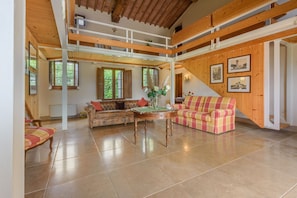 A spacious living area with a high ceiling, wooden beams, a sofa, a chair, and a table.
