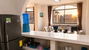A compact kitchen with a refrigerator, a dining table with chairs, a window with curtains, and a view of buildings. - Blue Horizon, Galápagos (San Cristóbal)