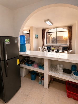A compact kitchen with a refrigerator, a dining table with chairs, a window with curtains, and a view of buildings.