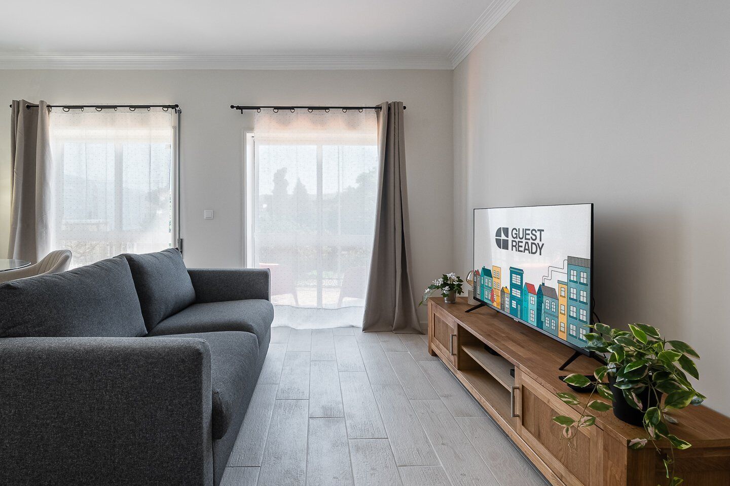 A modern living room with a grey sofa, a wooden TV stand with a 'GUEST READY' sign, and a large window with curtains.