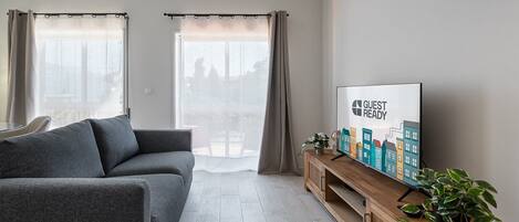 A modern living room with a grey sofa, a wooden TV stand with a 'GUEST READY' sign, and a large window with curtains.