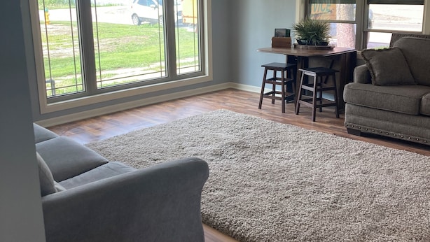 Amplia sala de estar con sofá gris, alfombra gris y piso de madera. Hay ventanas grandes con vistas al exterior.