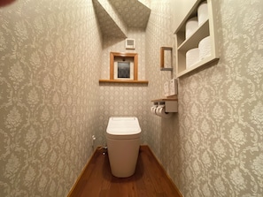 Une petite salle de bain avec des toilettes, un plancher en bois, un papier peint à motifs, une fenêtre et une étagère avec des serviettes.