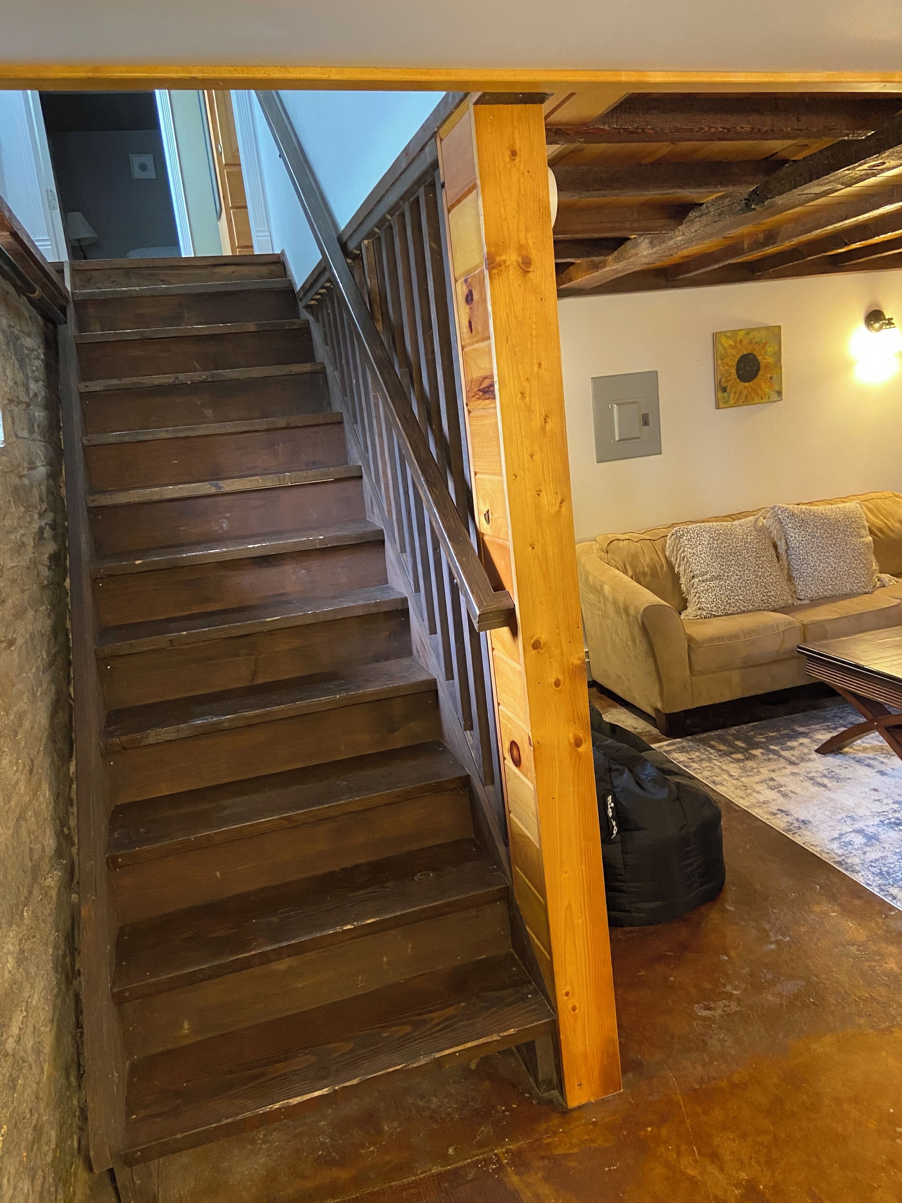 Un escalier en bois avec une rampe, menant à une pièce où se trouvent un canapé beige, un coussin décoratif à motifs et une petite table.