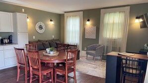A dining area with a wooden table and chairs, a kitchen with white cabinets, and a living area with a sofa and a decorative wall clock.
