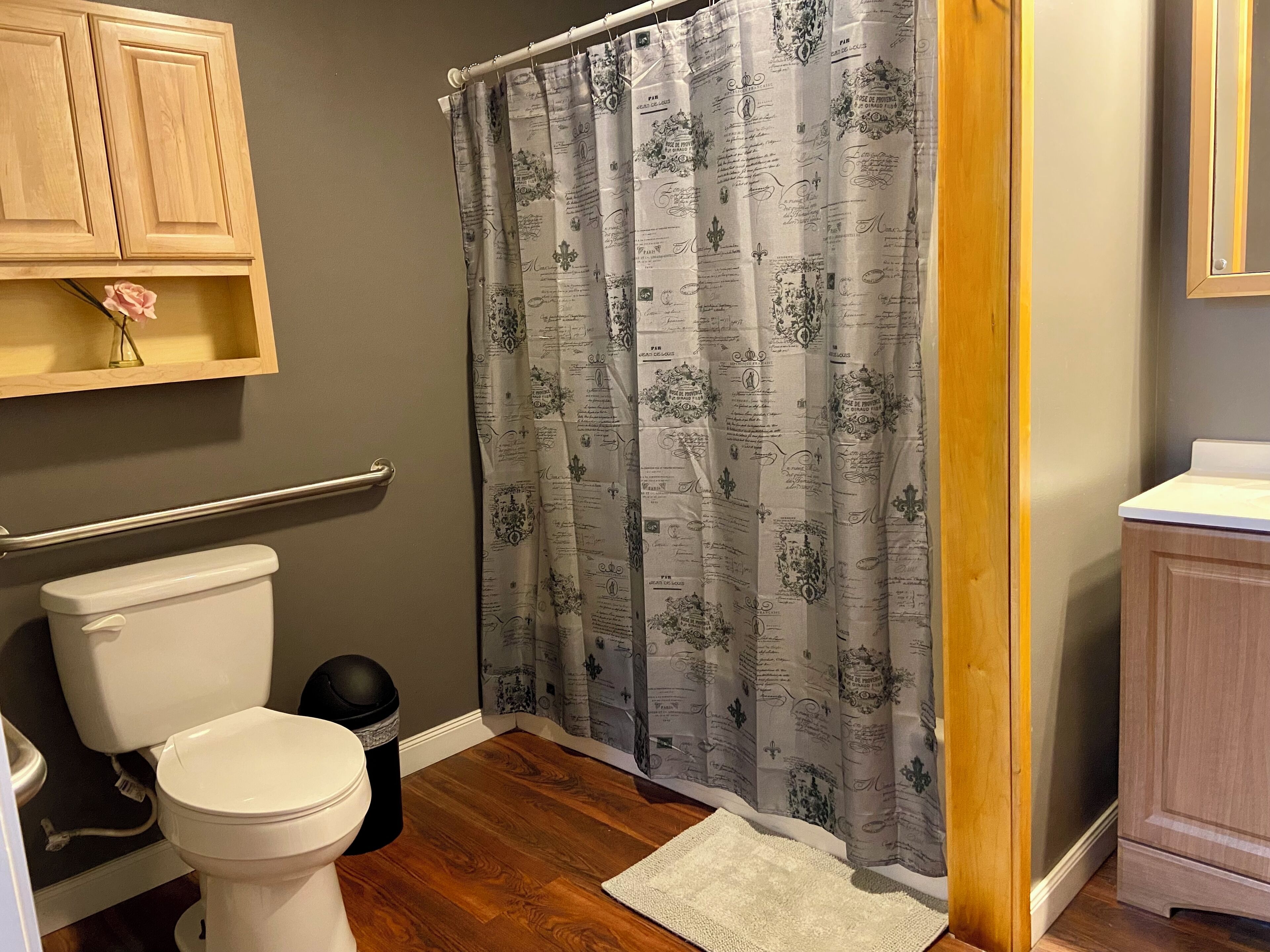 A bathroom with a toilet, a wooden cabinet, a shower curtain with a pattern, a white rug, and a wooden floor.