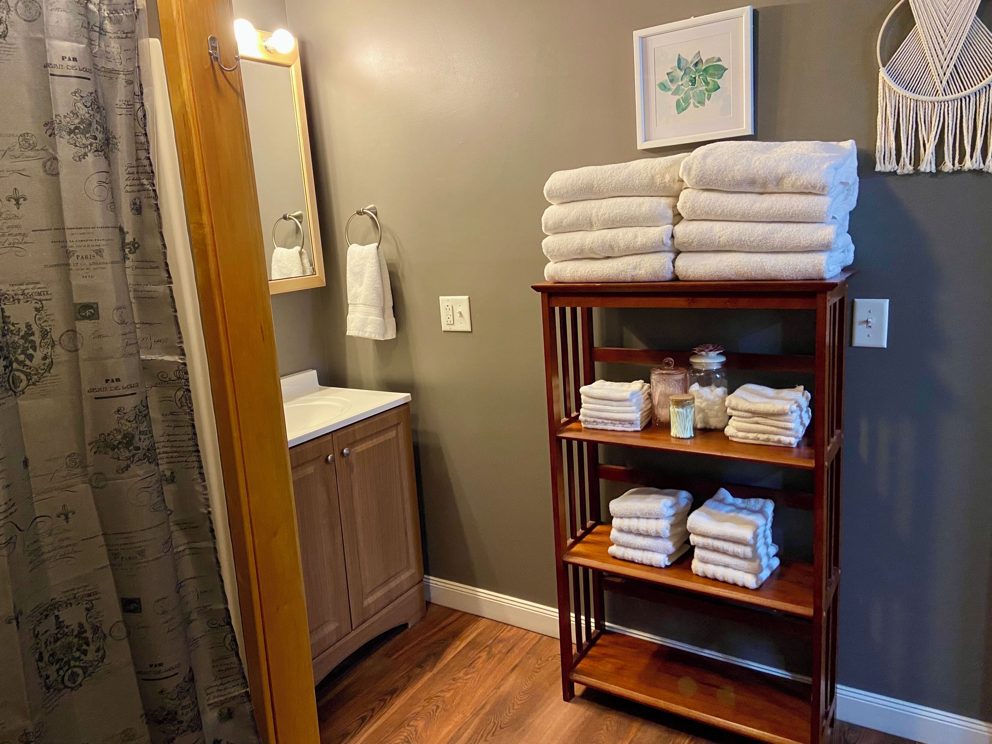 A bathroom with a wooden vanity, a mirror with lights, a towel rack, and a shelf with towels and toiletries.