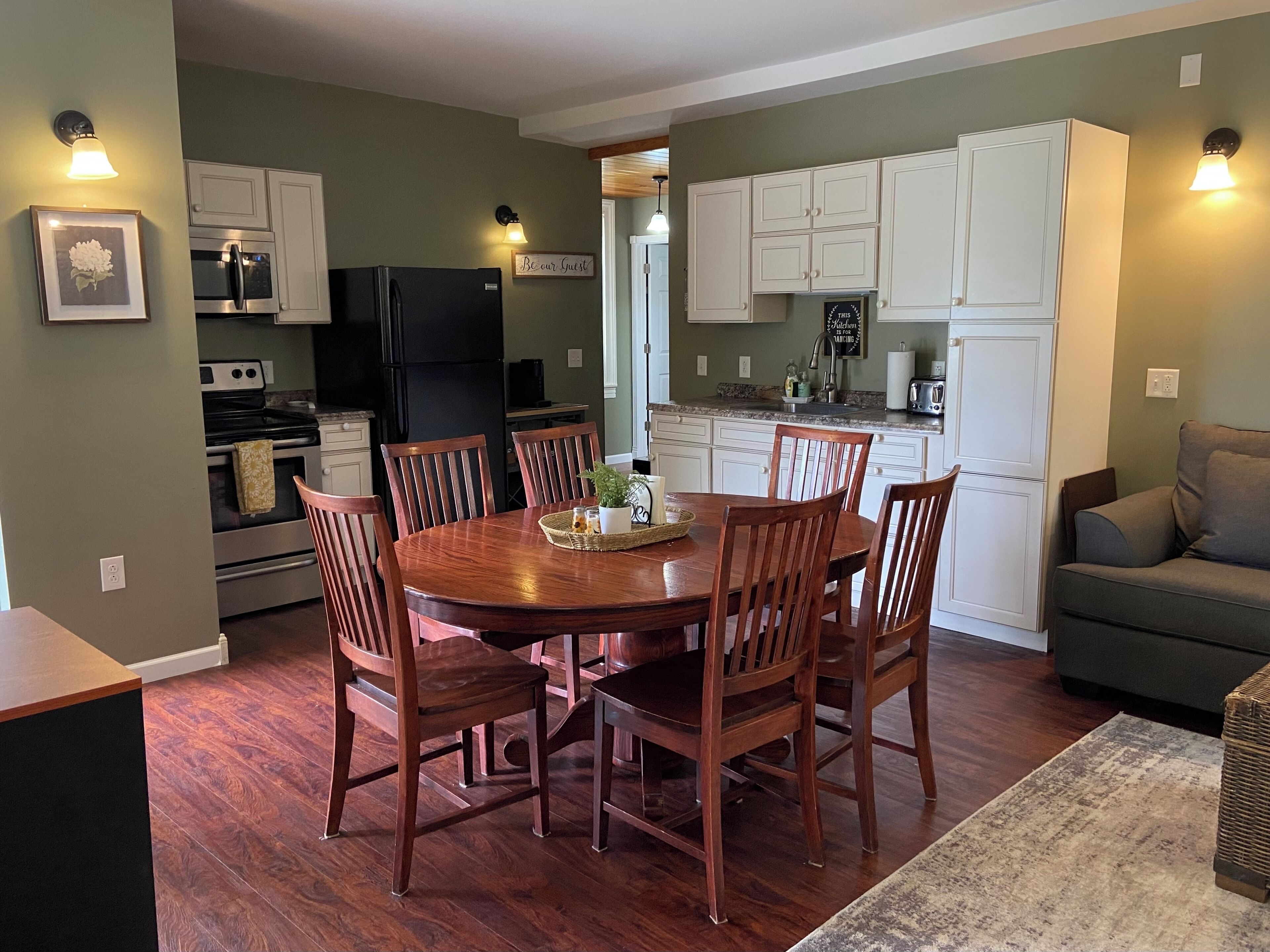 A dining area with a wooden table and chairs, a kitchen with white cabinets, a black refrigerator, and a gray sofa.