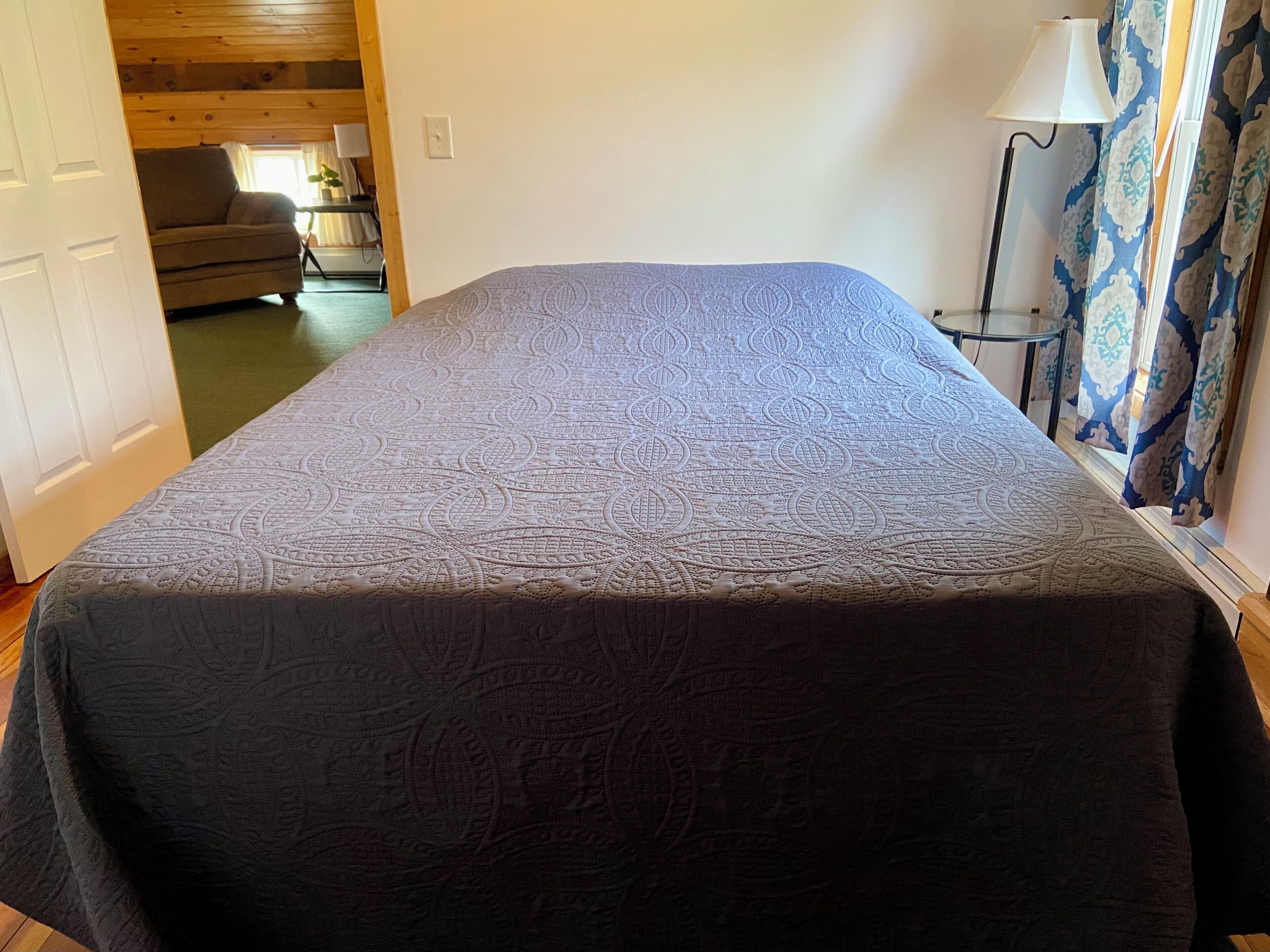 Ein Zimmer mit einem Bett, einem Sessel, einer Lampe und einem Fenster mit Vorhängen.