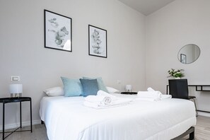 Een hotelkamer met een bed, twee kunstwerken in lijsten, een ronde spiegel, een bureau met een lamp, een stoel en een klein tafeltje met een bloevas.