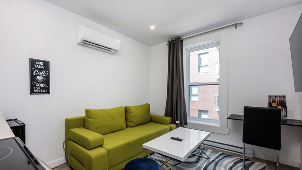 Une pièce moderne et bien éclairée, avec un canapé vert, une table blanche, un pouf bleu et une chaise noire. Un climatiseur est fixé au mur, et une fenêtre avec des rideaux est visible.