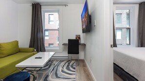Une chambre d’hôtel moderne avec un canapé vert, un bureau blanc, un téléviseur à écran plat, un lit et une vue sur la ville par les fenêtres.