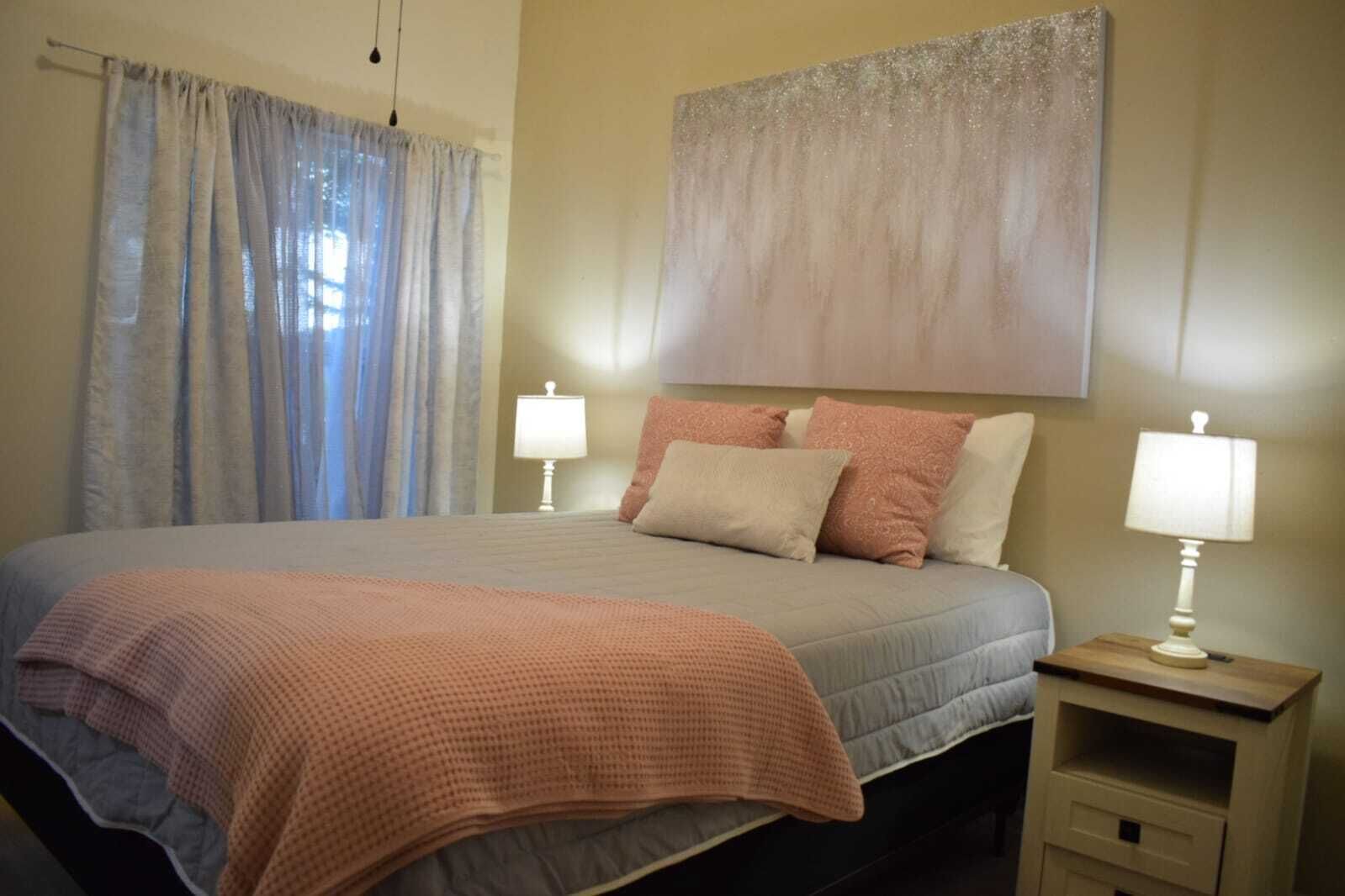 Un lit bien fait, recouvert d’une couverture rose, avec deux lampes de chevet et une grande peinture abstraite accrochée au mur.