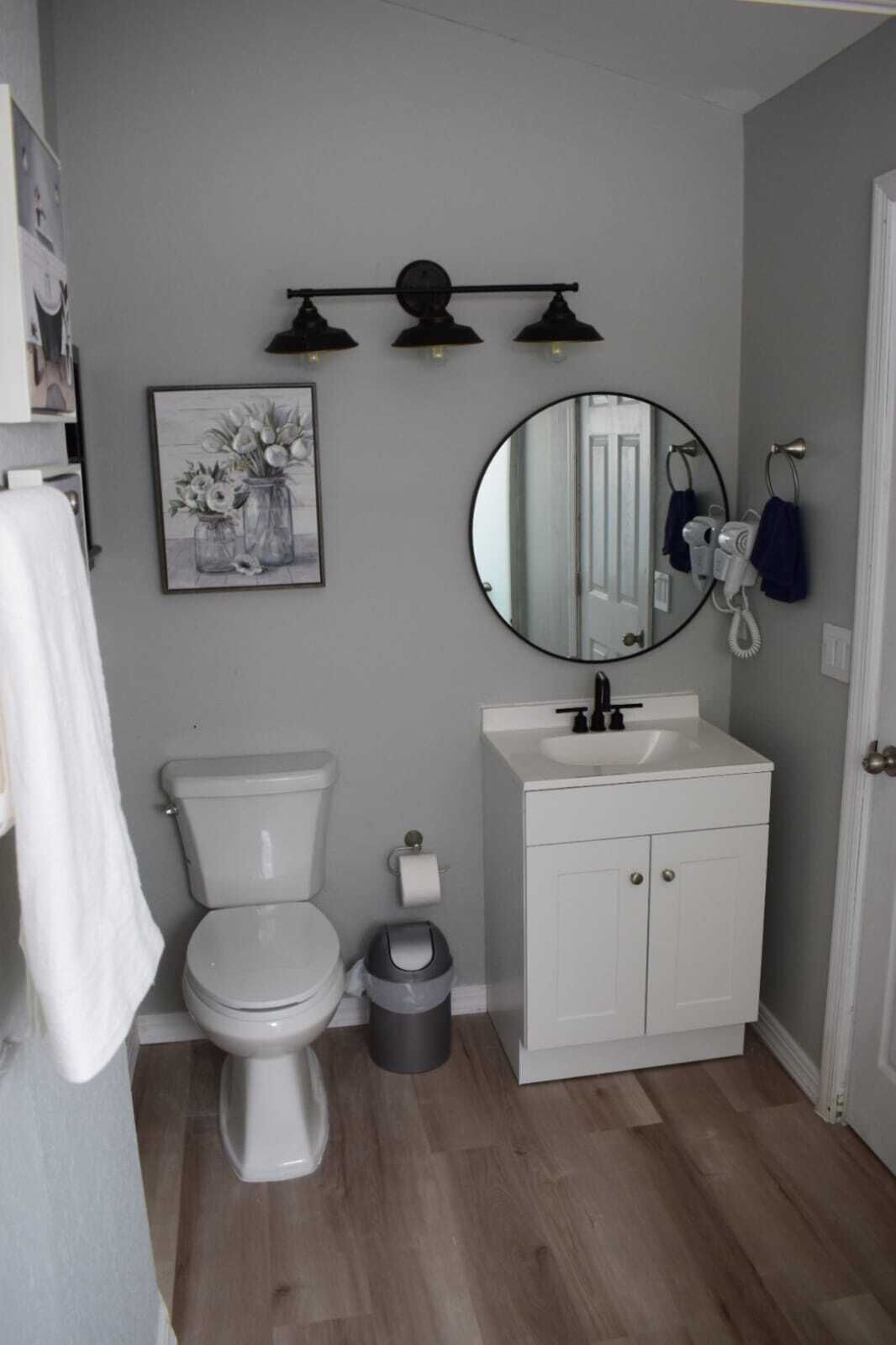 A bathroom with a toilet, sink, and mirror. There is a trash can and a towel rack.