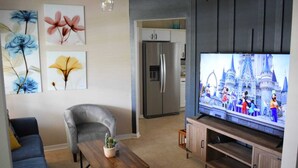 A living room with a grey armchair, a wooden coffee table with a cactus, a flat-screen TV displaying animated characters, and a kitchen with a refrigerator in the background. - Zojo_ Lovely House Close TO Disney (Clermont)
