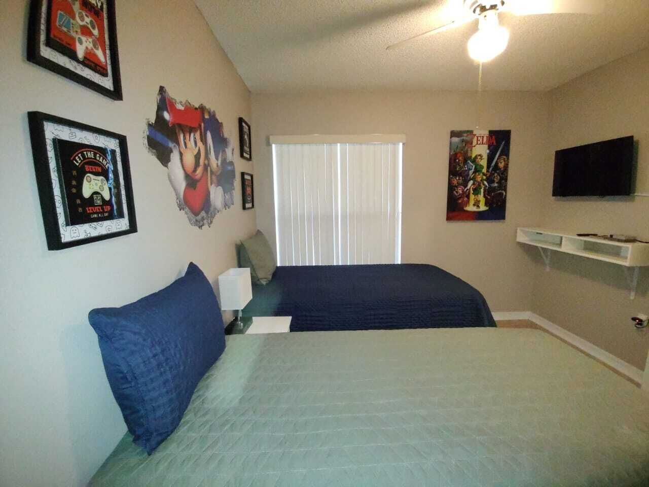 A bedroom with two beds, a window with blinds, a TV on a stand, and various posters on the walls.