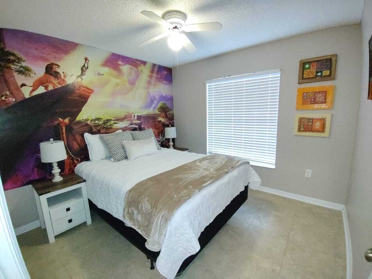 Un dormitorio con cama, mesitas de noche, ventilador de techo, ventana con persianas y un mural en la pared.
