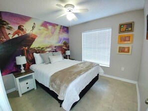 Un dormitorio con cama, mesitas de noche, ventilador de techo, ventana con persianas y un mural en la pared.