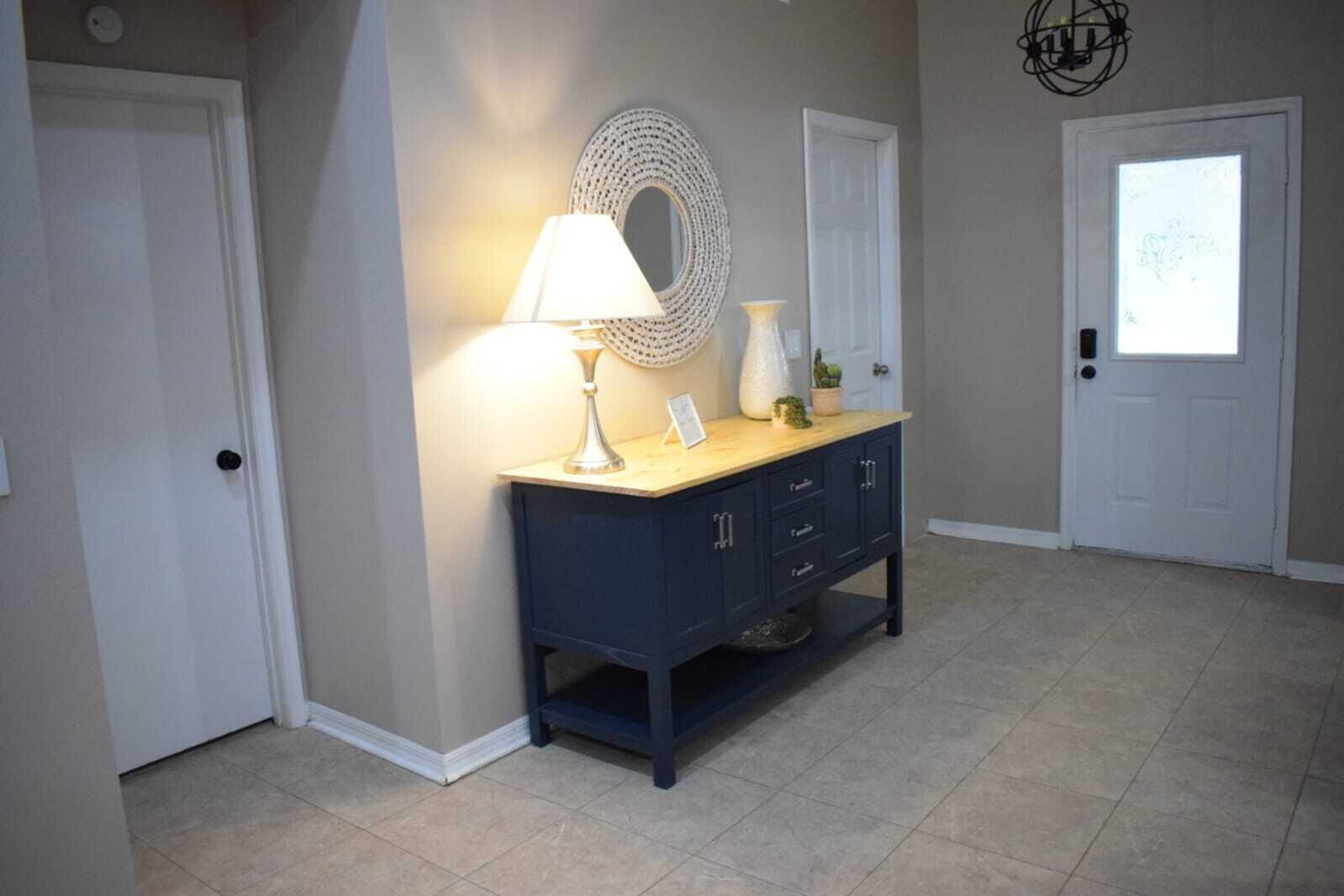 Un pasillo con una mesa consola azul oscuro, una lámpara blanca, un espejo decorativo y una puerta con vidrio esmerilado.