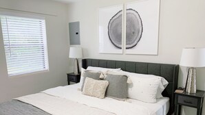Ein ordentlich bezogenes Bett mit einer grauen Decke, zwei Nachttische mit Lampen, ein Fenster mit Jalousien und zwei gerahmte Kunstwerke an der Wand.