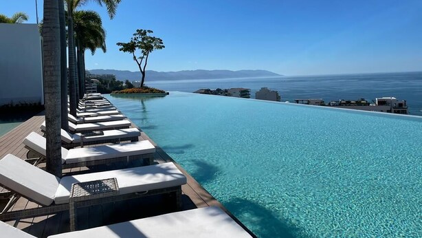 A infinity pool with lounge chairs and palm trees, overlooking a clear blue sea and distant mountains. - SOHO PV PUERTO VALLARTA  (Puerto Vallarta)