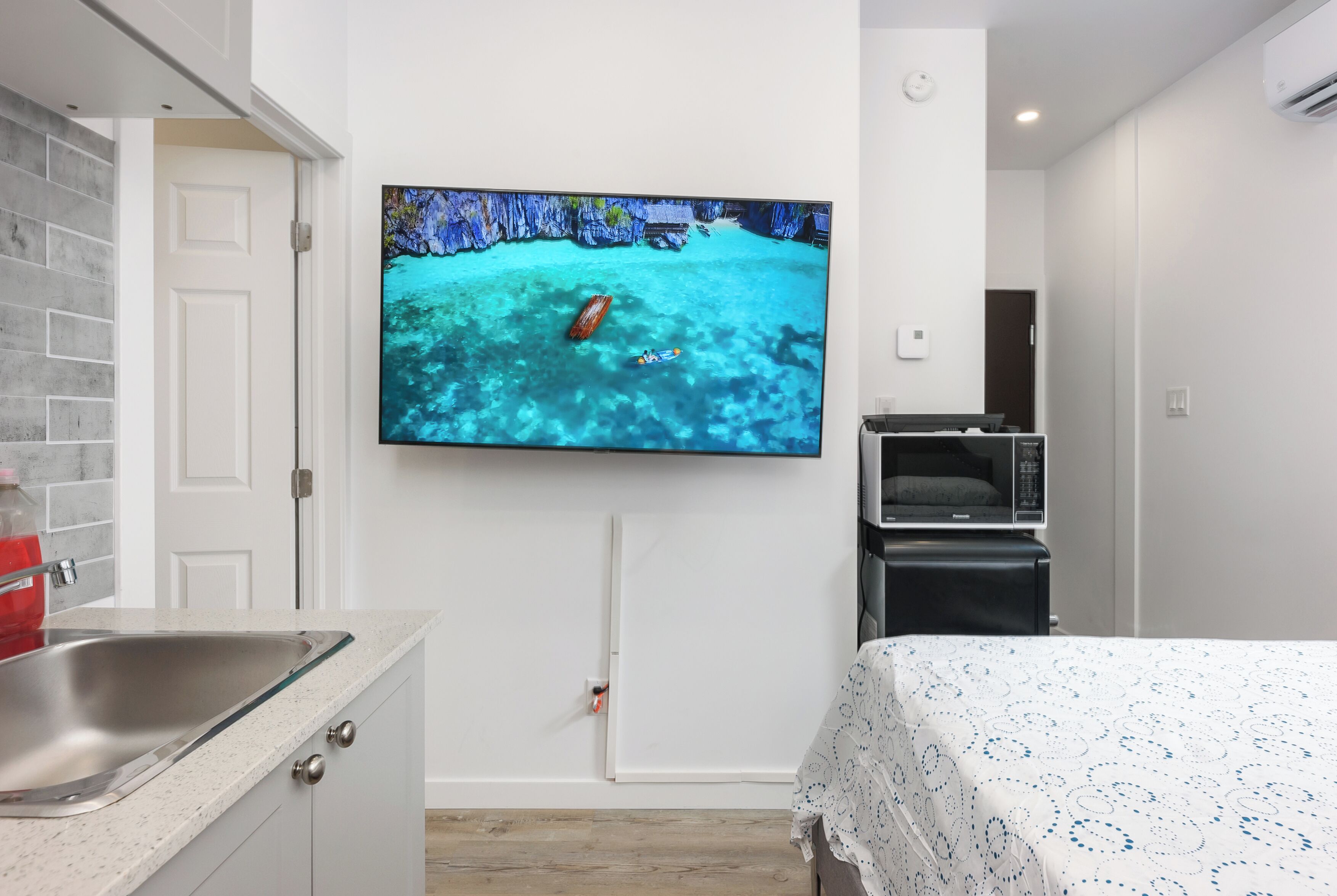 Ein modernes Hotelzimmer mit großem Flachbildfernseher an der Wand, einem Bett mit gemustertem Überwurf, einer kleinen Küchenzeile mit Mikrowelle und einem Badezimmer mit Duschvorhang.